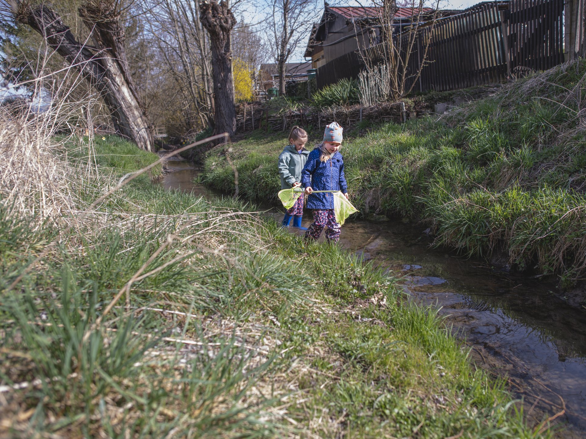 Zwei Kinder mit Gummistiefeln stehen am Ufer eines kleinen Bachs und halten einen Kescher ins Wasser. Um sie herum wachsen grüne Gräser und Bäume.