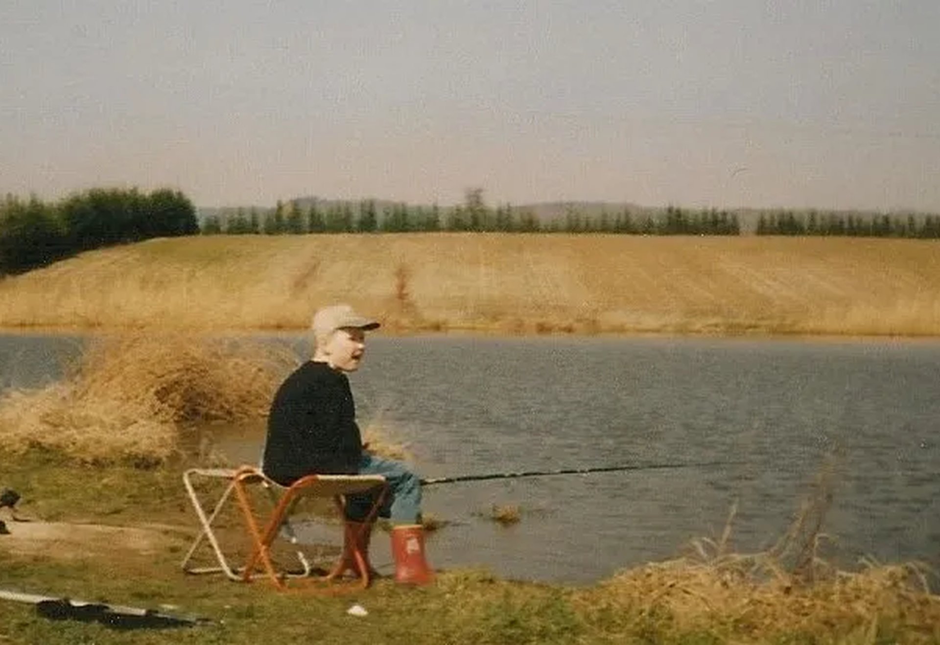 Ein Mann sitzt auf einem Klappstuhl am Ufer eines Sees und angelt, im Hintergrund Felder und Bäume.