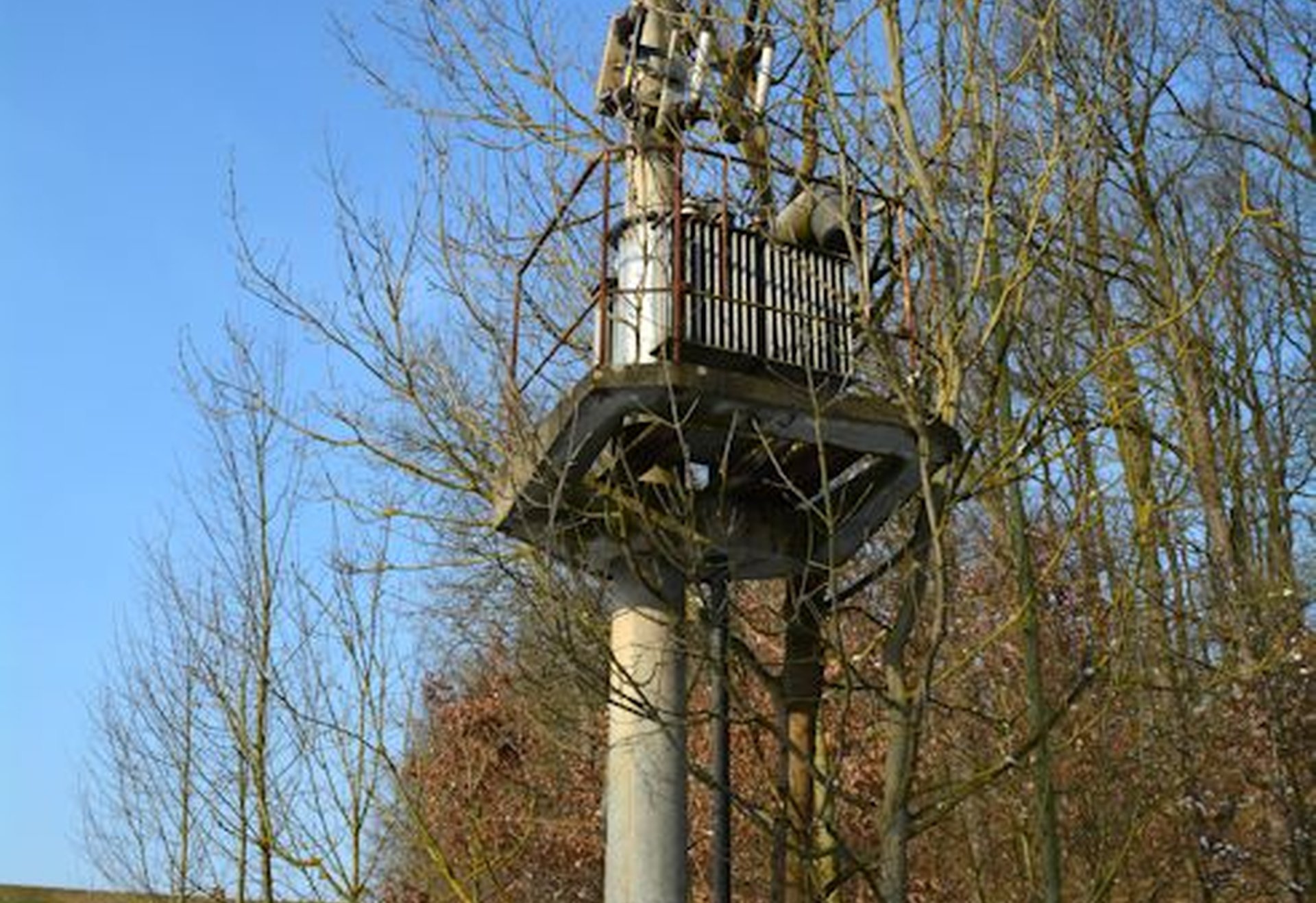 Ein altes, freistehendes Bahnsignal aus Metall auf einem Betonsockel steht zwischen kahlen Bäumen in einer winterlichen Landschaft.