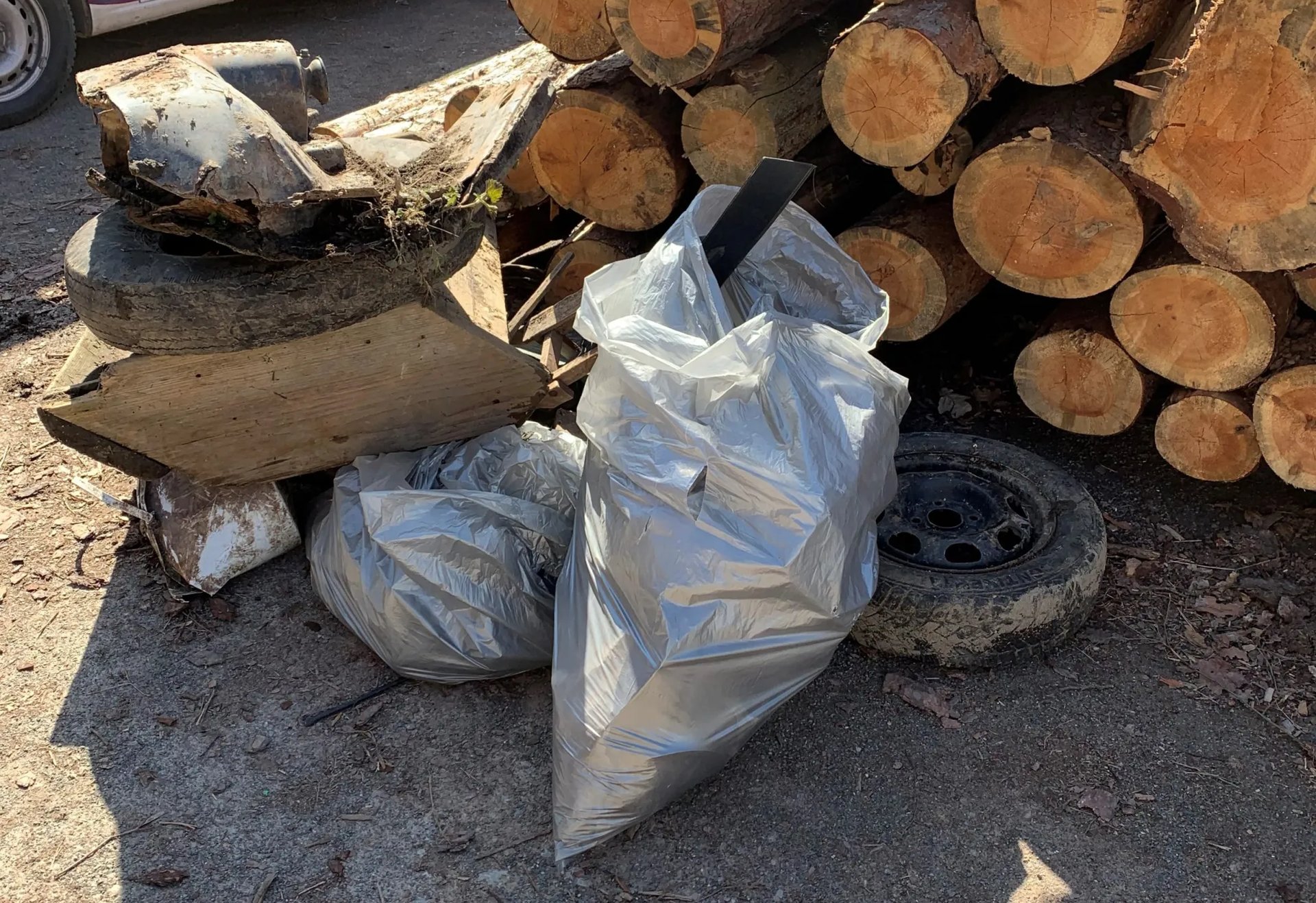 Zwei volle Müllsäcke, Autoteile und Holzstämme liegen auf einem Boden im Freien.