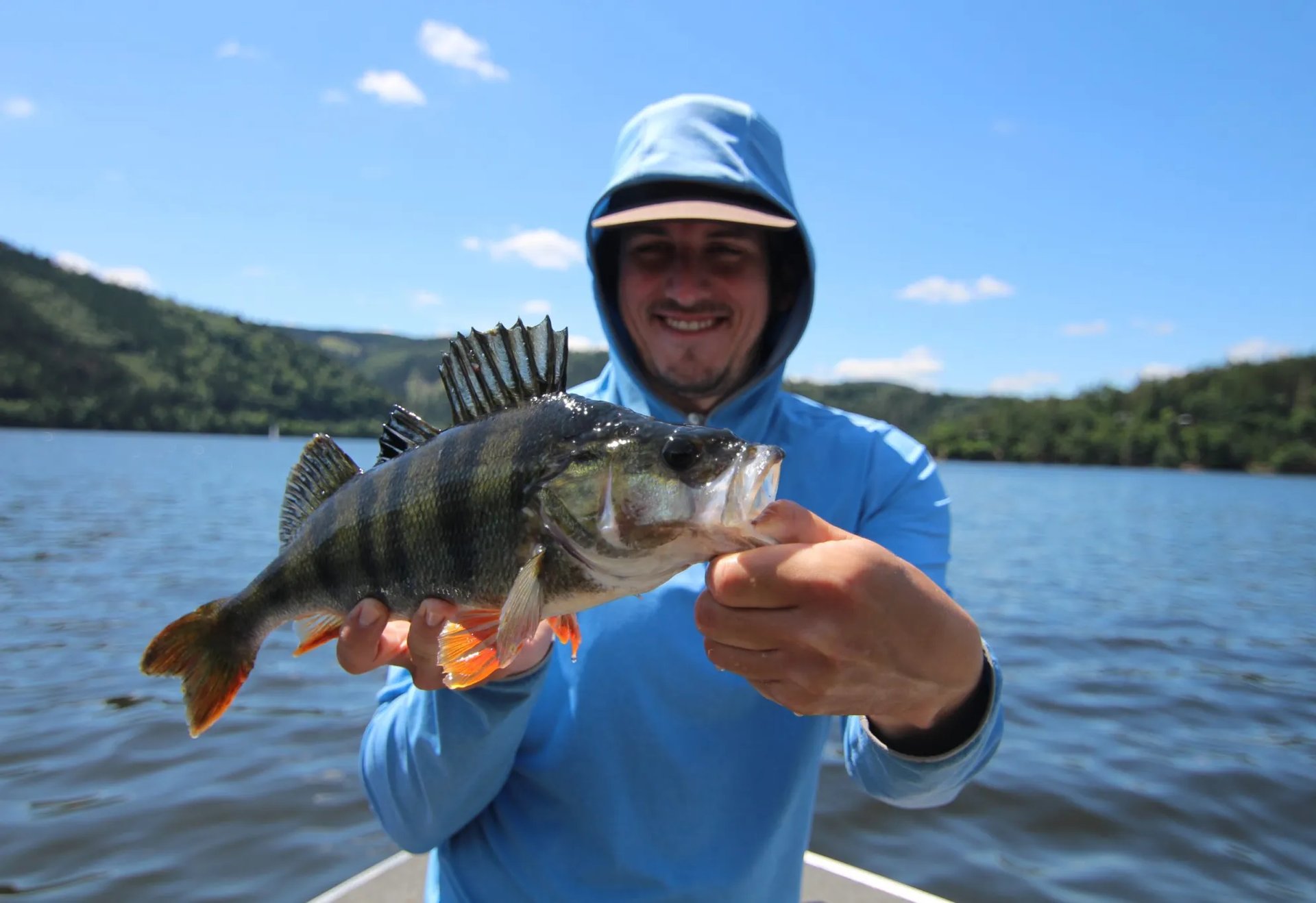 Ein Mann in blauer Kleidung hält lächelnd einen großen Fisch vor sich, im Hintergrund ein See und bewaldete Hügel.