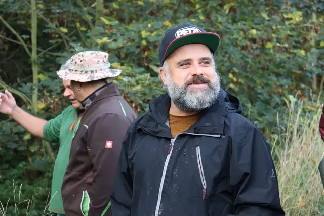 Zwei Männer stehen im Freien vor grünem Wald; der vordere Mann trägt Bart, Basecap und schwarze Jacke, der hintere einen Anglerhut und braune Jacke.