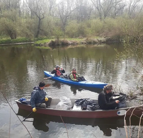 Drei Personen in zwei Booten sammeln Müll aus einem Fluss, umgeben von Bäumen und Gebüsch.