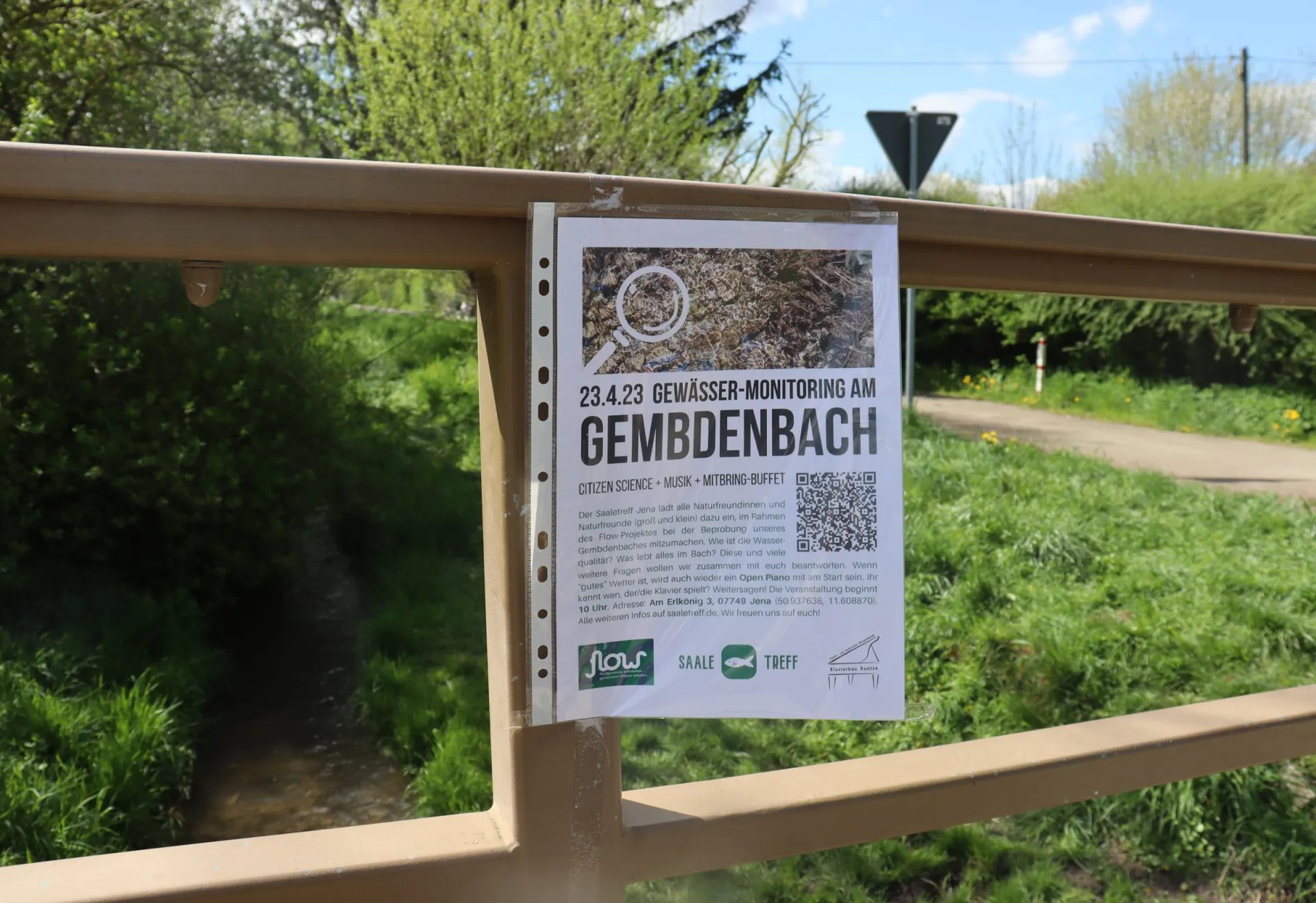Aushang mit Ankündigung zum "Gewässer-Monitoring am Gembdenbach" an einem Brückengeländer in grüner, ländlicher Umgebung.