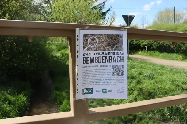 Aushang mit Ankündigung zum "Gewässer-Monitoring am Gembdenbach" an einem Brückengeländer in grüner, ländlicher Umgebung.