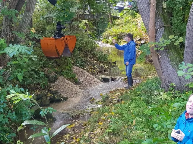 Eine Person steht an einem kleinen Bach und zeigt auf einen Baggerlöffel, der Kies ins Wasser schüttet; im Vordergrund steht eine weitere Person.