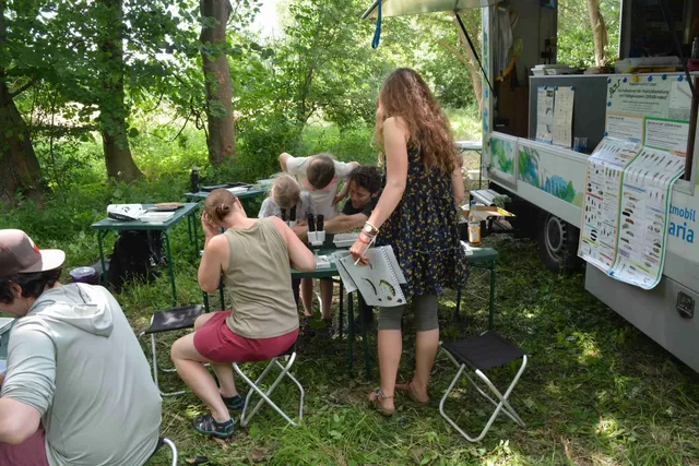 Mehrere Menschen arbeiten und forschen gemeinsam an Tischen im Freien neben einem mit Infopostern ausgestatteten Fahrzeug in einem grünen, bewaldeten Gebiet.