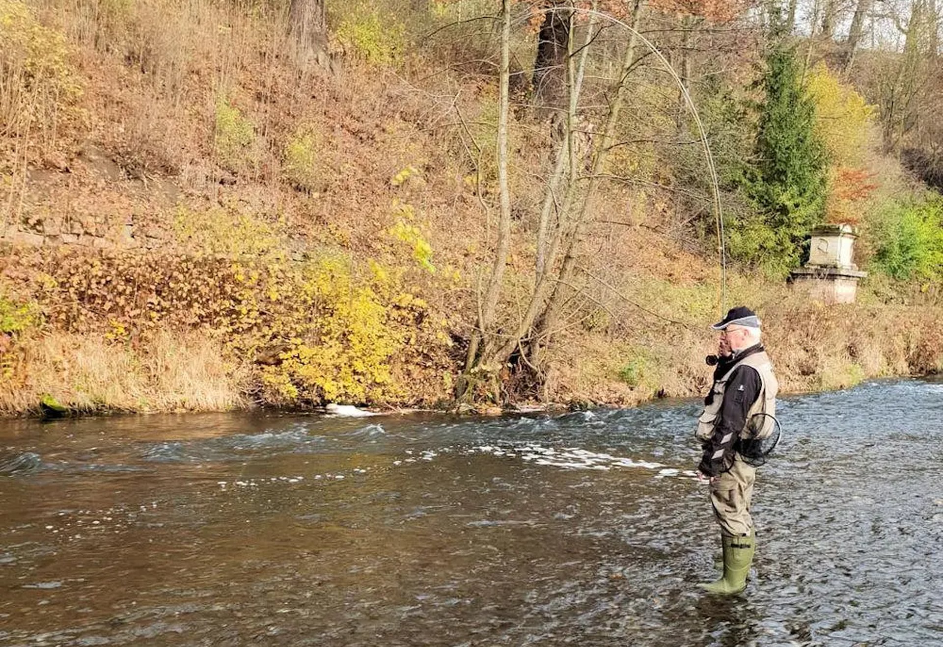 Ein Mann mit Angelkleidung steht in einem flachen Fluss und angelt.