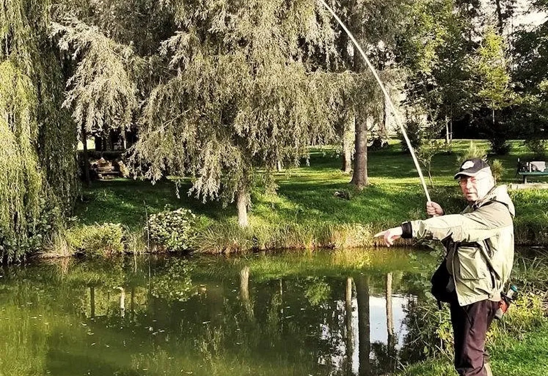 Ein Angler steht am Ufer eines kleinen Teichs und hält eine stark gebogene Angelrute ins Wasser.
