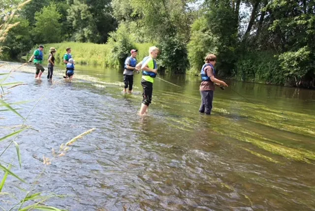 Mehrere Personen, darunter Kinder und Erwachsene, waten mit Schwimmwesten durch einen flachen Fluss in einer grünen, bewaldeten Umgebung.
