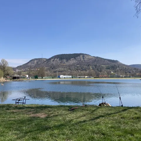 Blick auf einen See mit Angelruten am Ufer, im Hintergrund ein bewaldeter Hügel unter klarem Himmel.