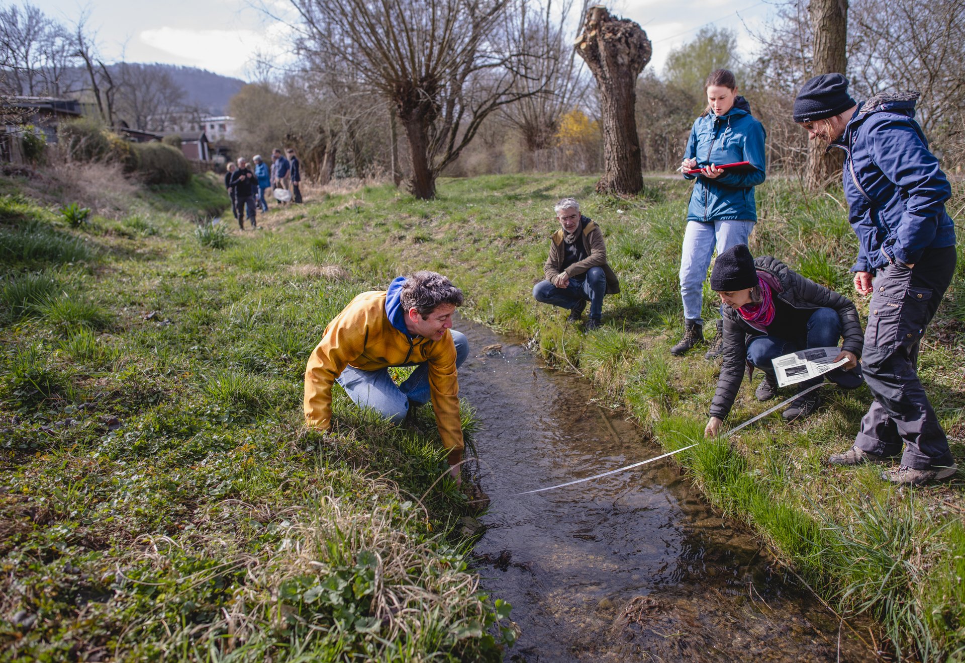 Mehrere Personen führen Messungen und Untersuchungen an einem kleinen Bach in einer naturnahen Wiese durch.
