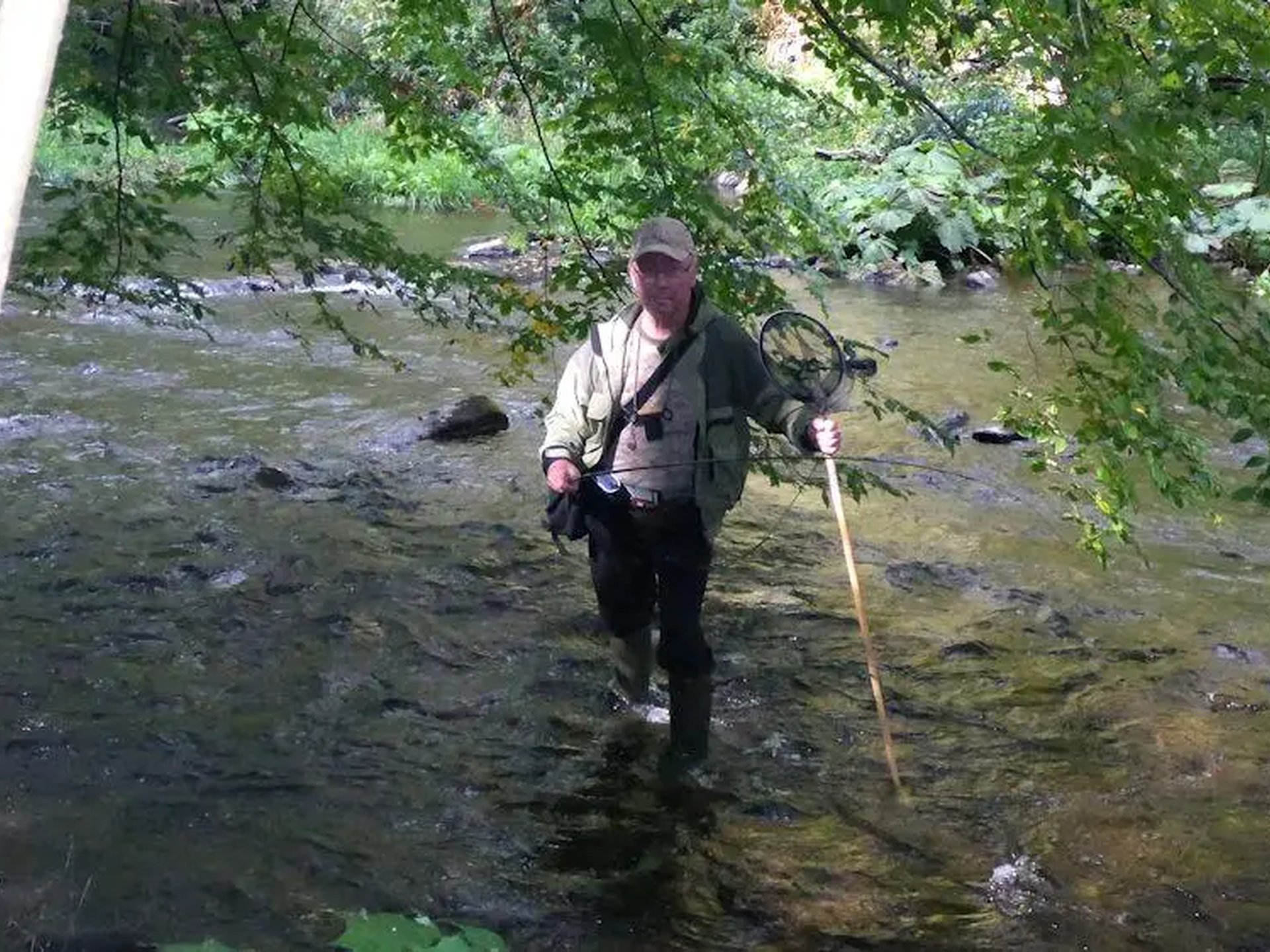Ein Mann steht mit Angelzubehör und einem Stock in einem flachen Fluss, umgeben von grüner Vegetation.