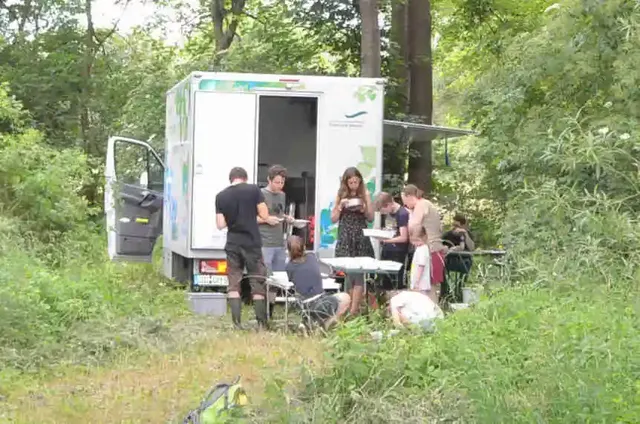 Eine Gruppe von Menschen steht und sitzt an Tischen vor einem weißen Versorgungsfahrzeug im Grünen.