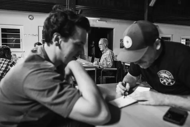 Zwei Männer sitzen an einem Tisch und schreiben gemeinsam auf ein Blatt Papier, im Hintergrund sitzen weitere Personen in einem Raum.