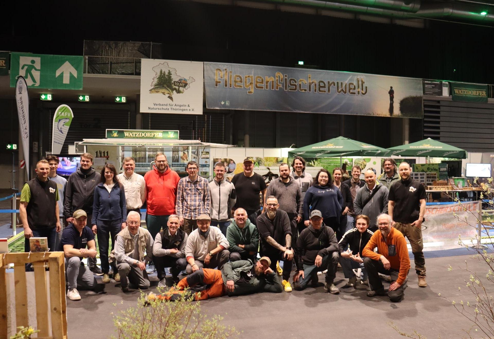 Eine Gruppe von etwa 25 Menschen steht und kniet zusammen in einer Messehalle vor Messeständen, über ihnen hängt ein Banner mit der Aufschrift „Fliegenfischerwelt“.
