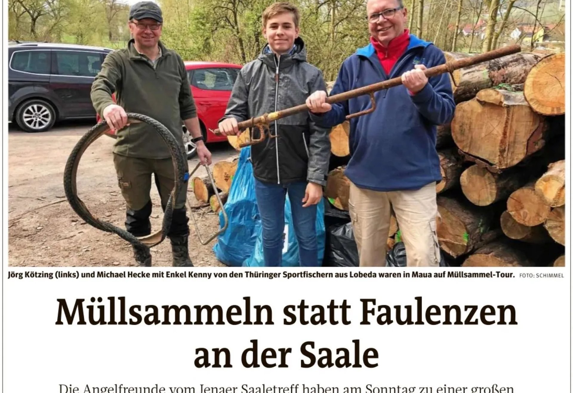 Drei Personen stehen lachend nebeneinander und halten gesammelten Müll, darunter ein altes Fahrradteil und einen großen Stock, im Hintergrund liegt gestapeltes Holz.