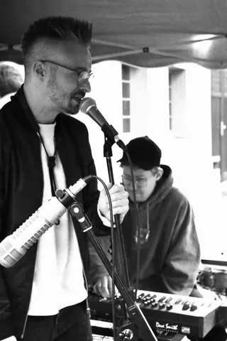 Ein Mann singt in ein Mikrofon, während eine zweite Person an einem Mischpult arbeitet.