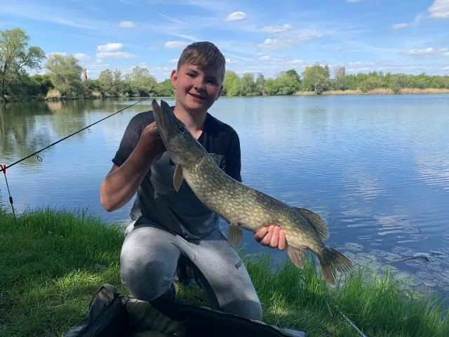 Ein Junge sitzt am Ufer eines Sees und hält lächelnd einen großen gefangenen Fisch in den Händen.