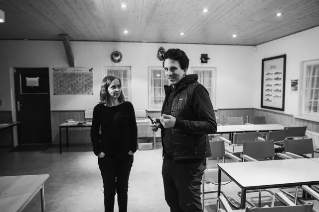 Zwei Personen stehen in einem leeren, hell beleuchteten Unterrichtsraum und unterhalten sich.