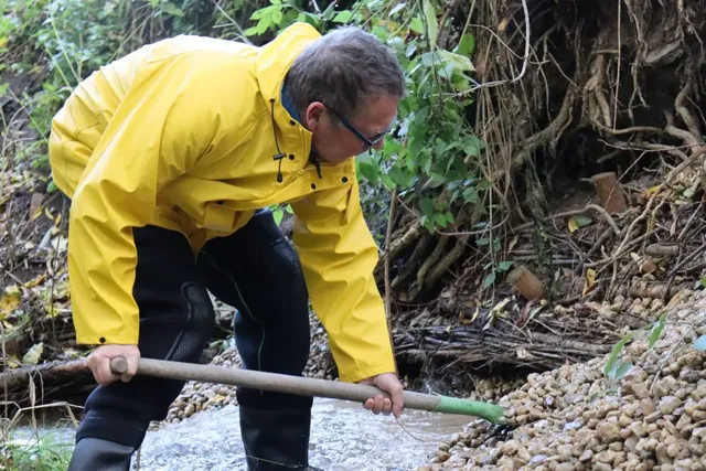 Ein Mann in gelber Regenjacke arbeitet mit einer Schaufel an einem kleinen Bach.