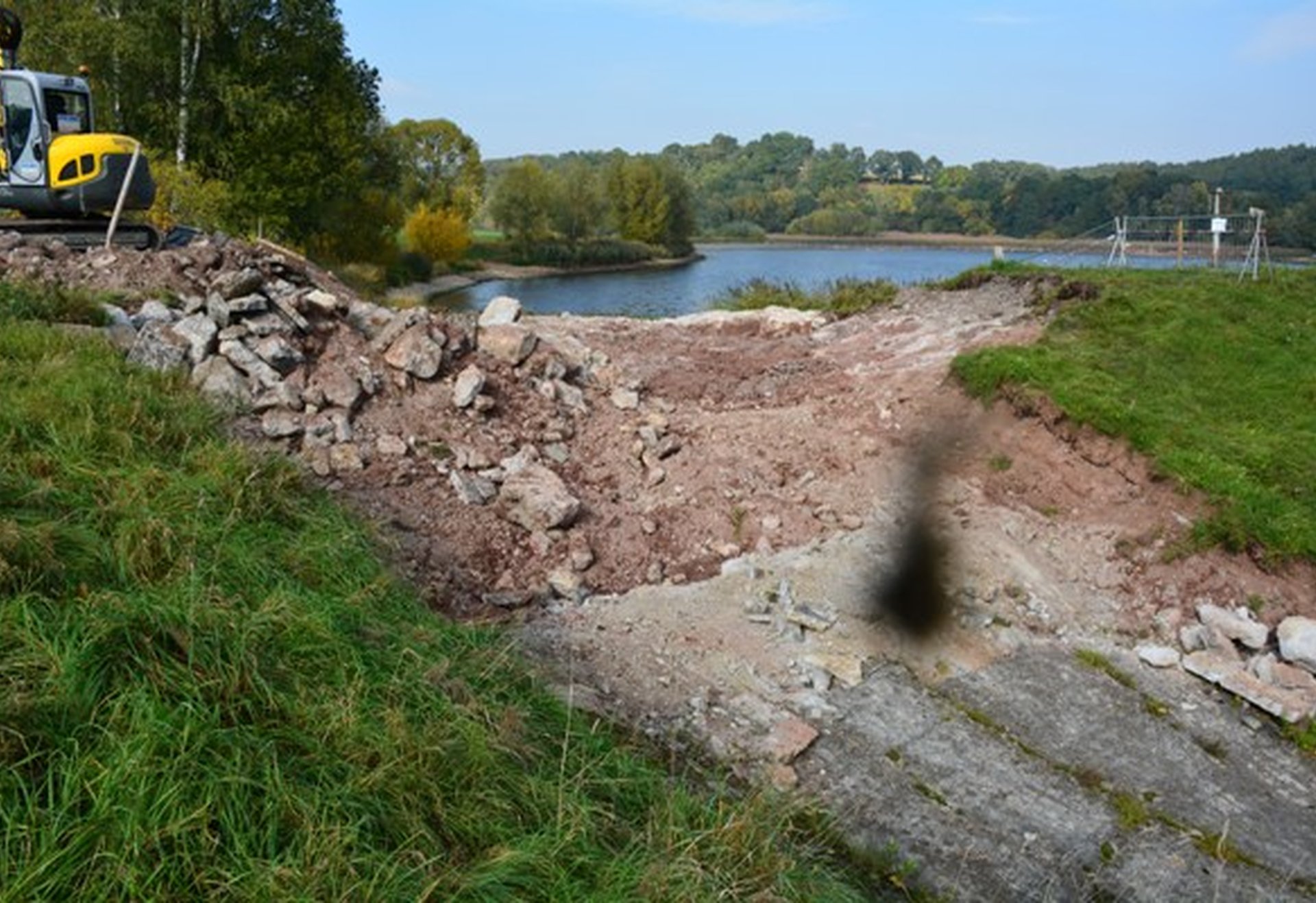 Ein zerstörter Damm mit aufgebrochenem Erdreich und Bagger, im Hintergrund ein See und Bäume.