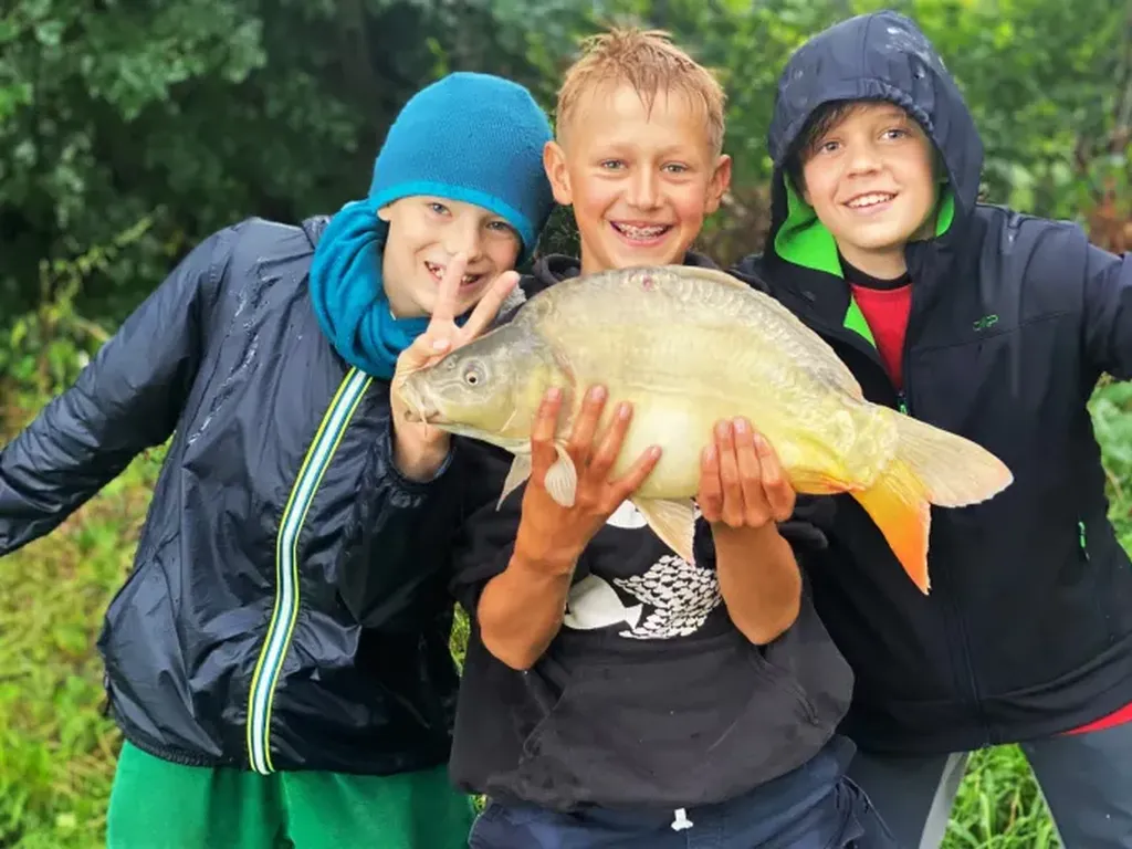 Drei lachende Kinder posieren im Freien, eins von ihnen hält einen großen Fisch in den Händen, die anderen beiden machen fröhliche Gesten.