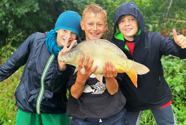 Drei lachende Kinder posieren im Freien, eins von ihnen hält einen großen Fisch in den Händen, die anderen beiden machen fröhliche Gesten.