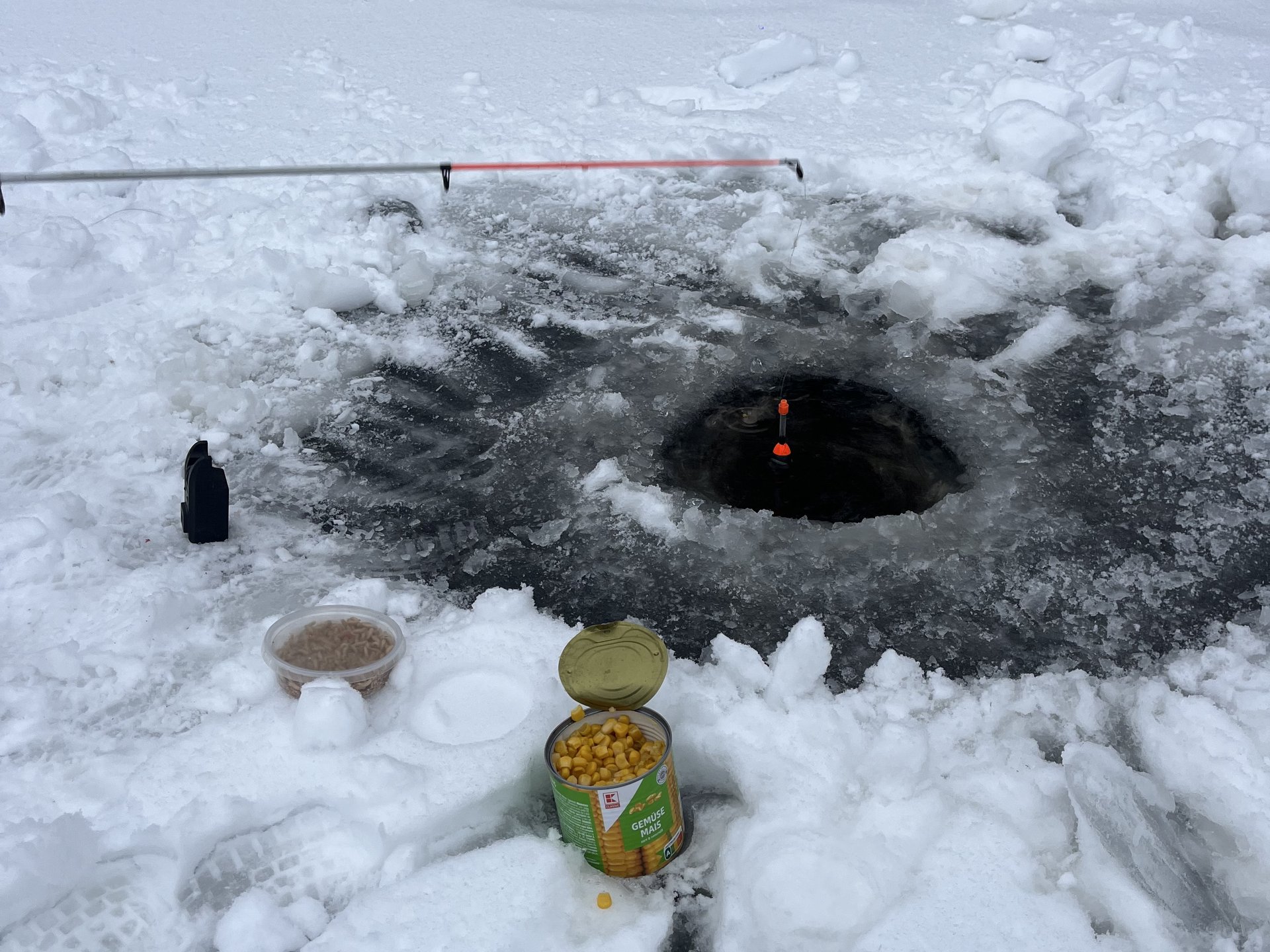 Angelrute mit Pose über einem Eisloch auf verschneitem See.