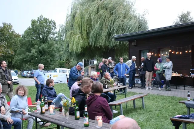 Menschen sitzen und stehen bei einem gemütlichen Gartenfest mit Getränken und Kerzen an Holztischen, im Hintergrund Bäume und ein Gebäude.