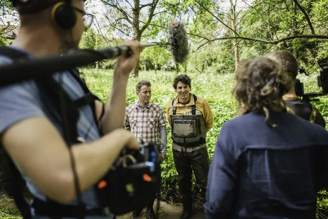 Ein Kamerateam filmt zwei Männer im Grünen, einer trägt Outdoor-Kleidung und einen Watoverall.