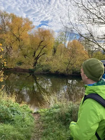 Eine Person in grüner Jacke steht am Ufer eines Flusses und schaut auf herbstliche Bäume.