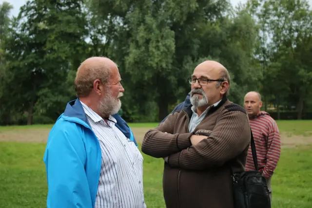 Zwei ältere Männer unterhalten sich im Freien, ein dritter Mann steht im Hintergrund auf einer Wiese.