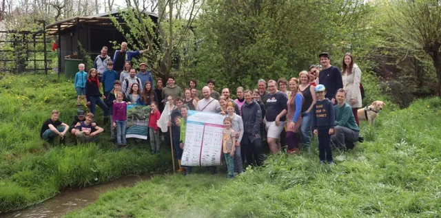 Eine große Gruppe von Menschen steht und sitzt auf einer Wiese im Grünen, einige halten Plakate, im Hintergrund sind Bäume und ein Schuppen zu sehen.