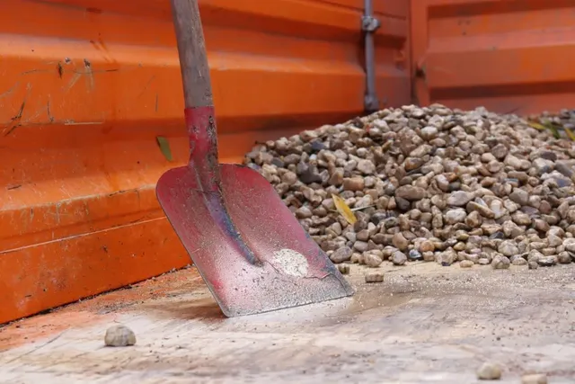Eine Schaufel lehnt an einem Haufen Kies in einem orangefarbenen Container.