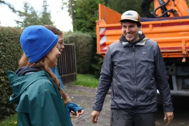Drei Personen unterhalten sich lachend im Freien vor einem orangefarbenen Lkw.