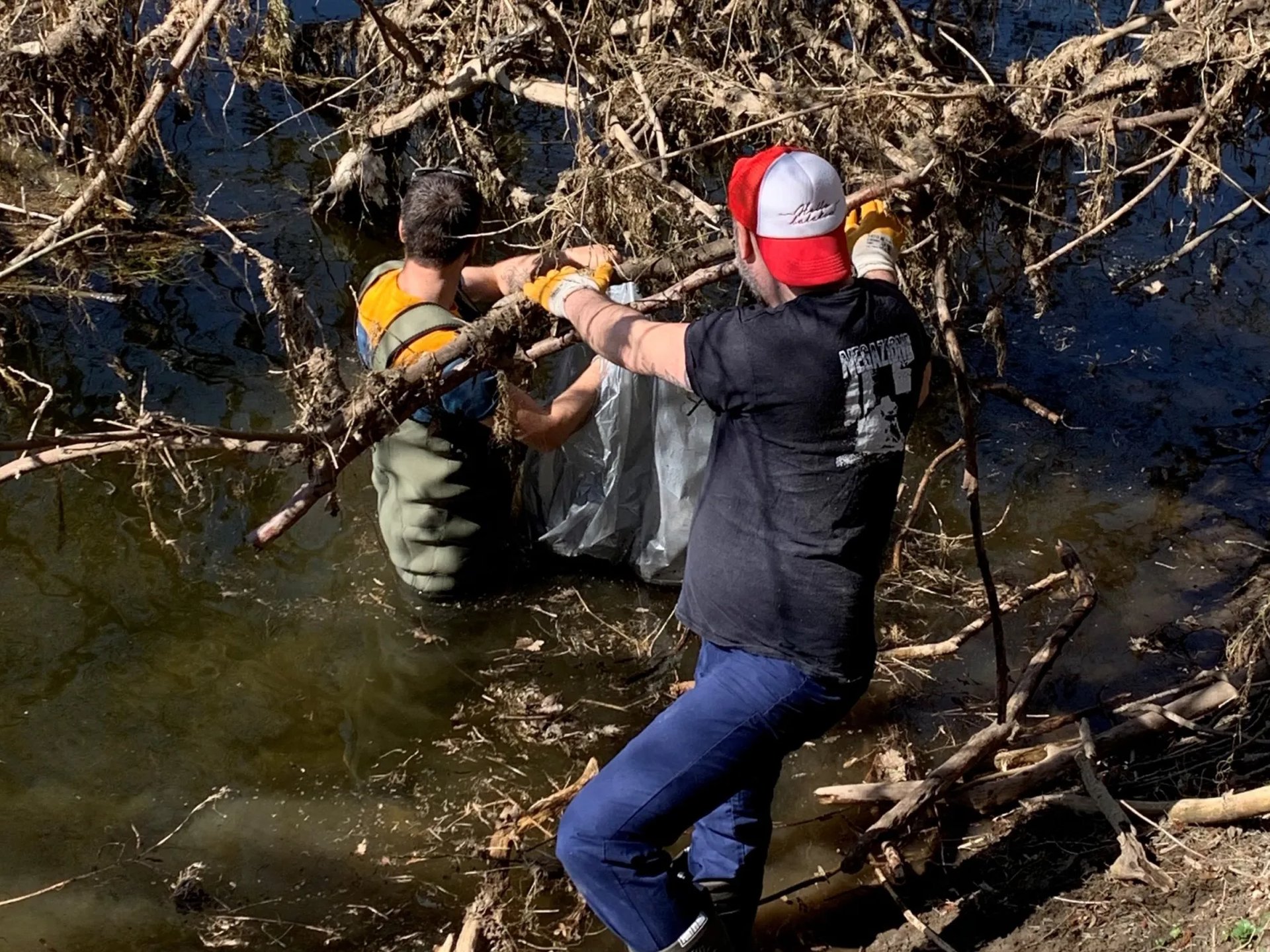 Zwei Männer entfernen mit Handschuhen und Werkzeugen Müll und Äste aus einem Fluss.