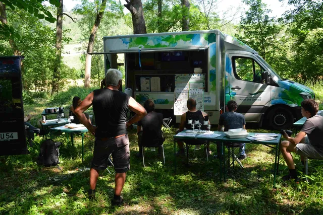 Mehrere Menschen sitzen an Tischen im Freien vor einem bunten Informationsbus im Wald und hören einer Person zu.