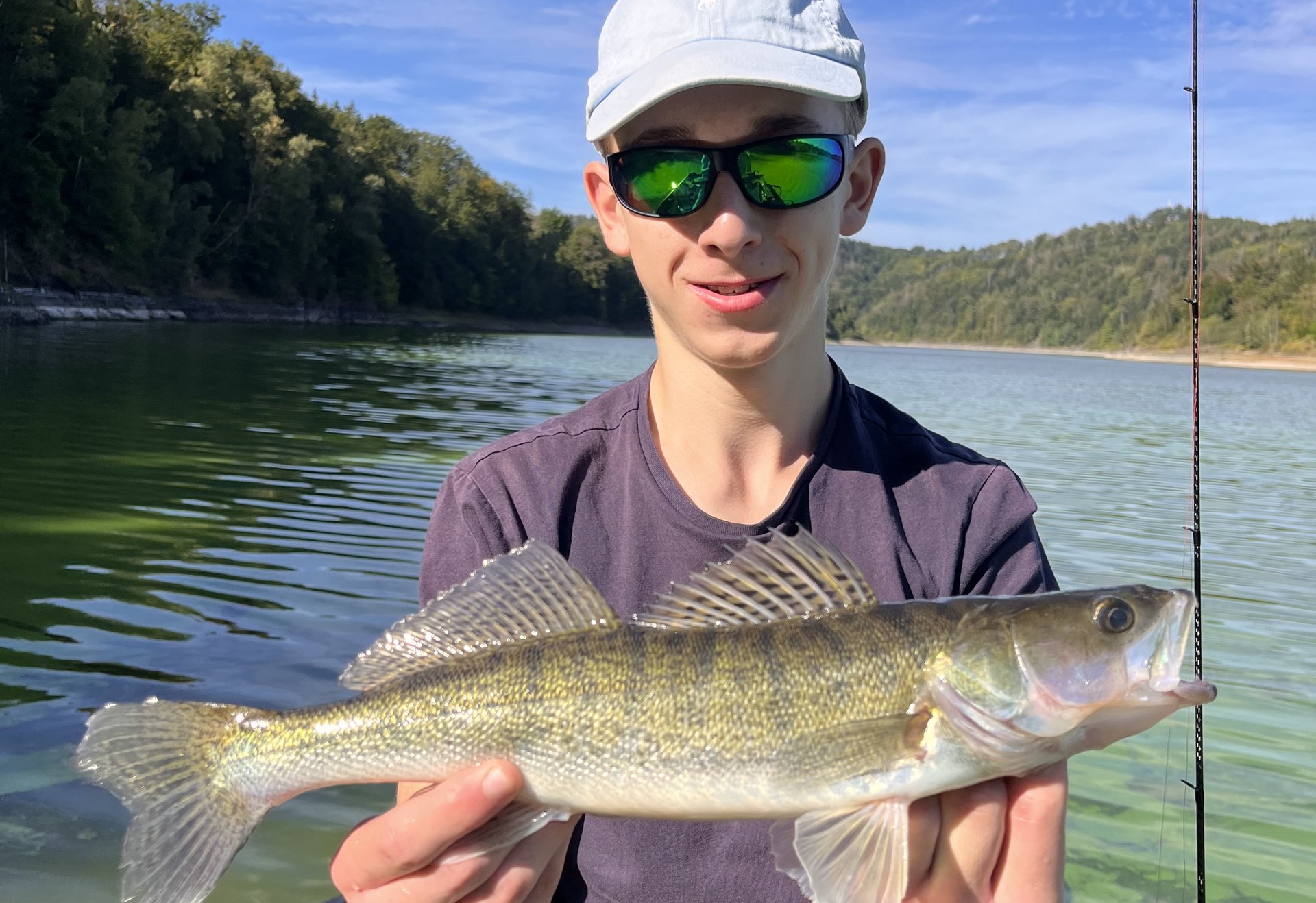 Ein junger Angler hält stolz einen frisch gefangenen Zander auf einem Boot. Im Hintergrund spiegeln sich bewaldete Ufer im klaren Wasser unter blauem Himmel.