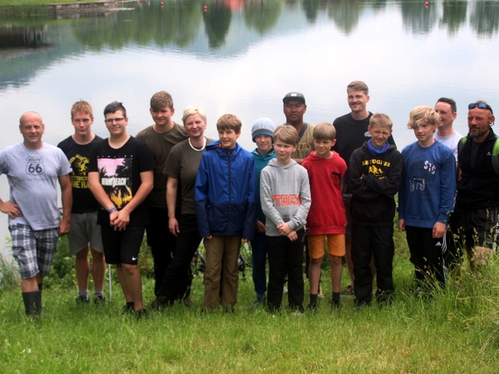 Eine Gruppe von Jugendlichen und Erwachsenen steht am Ufer eines Sees auf einer Wiese und blickt in die Kamera.