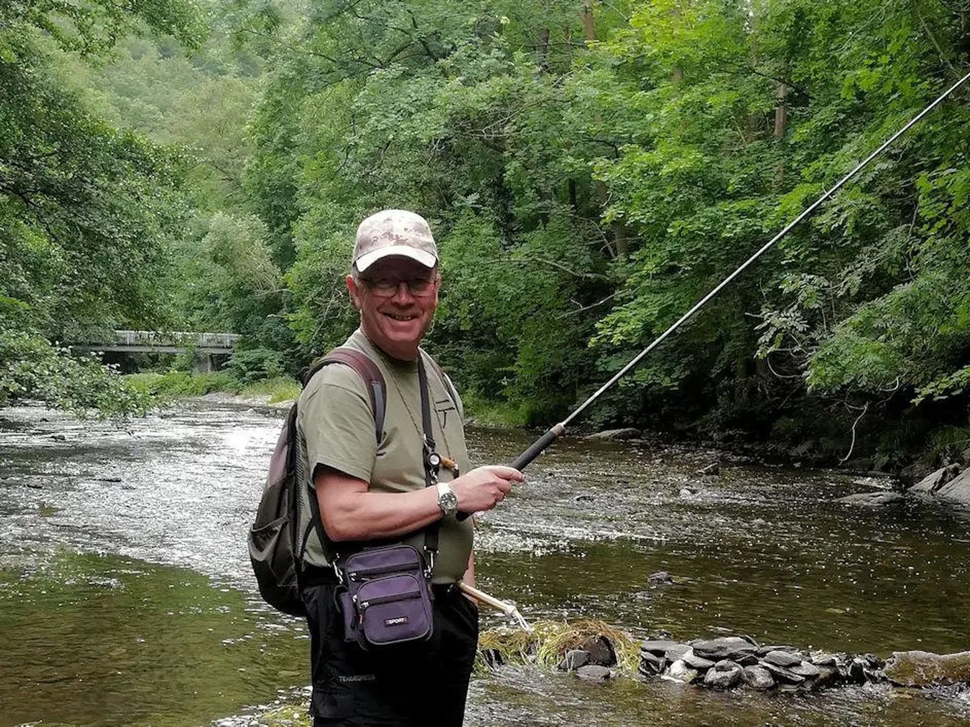 Ein Mann angelt mit einer Angelrute an einem Fluss, umgeben von grüner Natur.