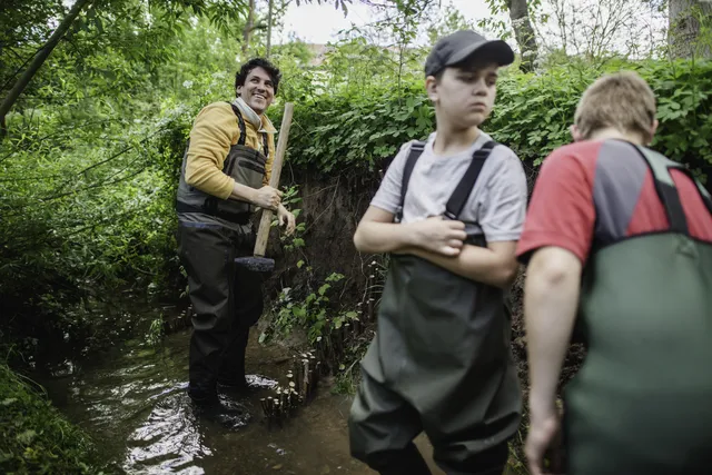 Drei Personen in Watthosen stehen in einem kleinen, bewaldeten Bach und arbeiten gemeinsam an einem Naturprojekt.