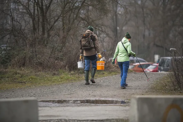 Zwei Personen gehen auf einem Waldweg spazieren, eine trägt Eimer und die andere sammelt mit einer Greifzange Müll auf.