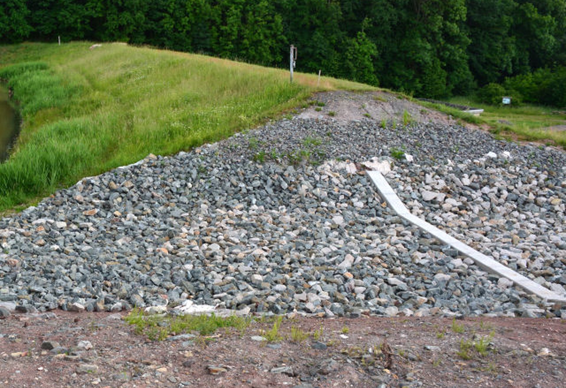 Böschung mit einer Steinschüttung, daneben eine Wiese und im Hintergrund Bäume.