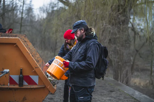 Zwei Personen entsorgen Müll in einen großen Container im Freien; eine trägt eine orangefarbene Eimer, die andere eine rote Mütze. Im Hintergrund sind Bäume und ein trüber Himmel zu sehen.