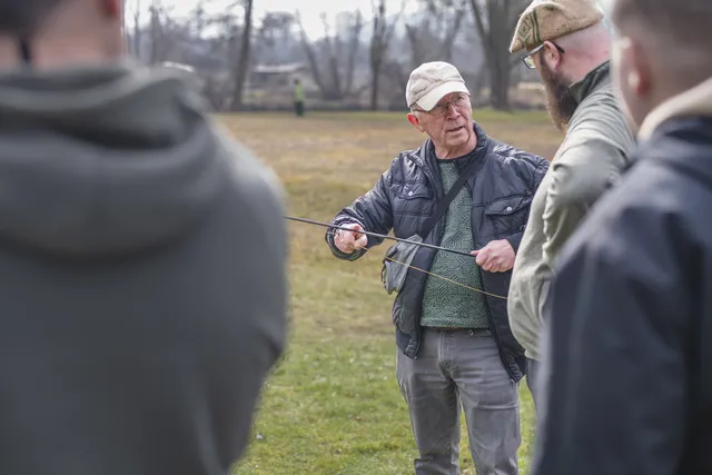 Ein älterer Mann erklärt einer kleinen Gruppe den Umgang mit einer Fliegenrute. Alle hören aufmerksam zu, die Stimmung ist konzentriert und freundlich.