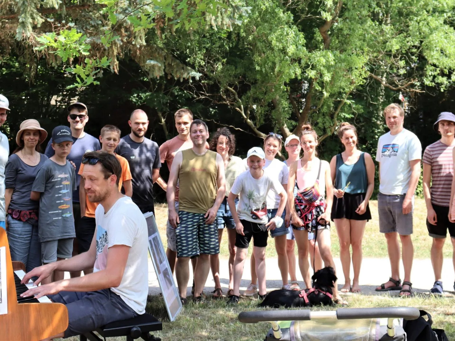 Eine Gruppe von Menschen steht im Freien im Schatten, während vorne ein Mann Klavier spielt.