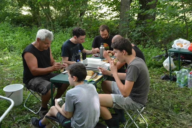 Sechs Personen sitzen an einem Tisch im Grünen und essen gemeinsam.