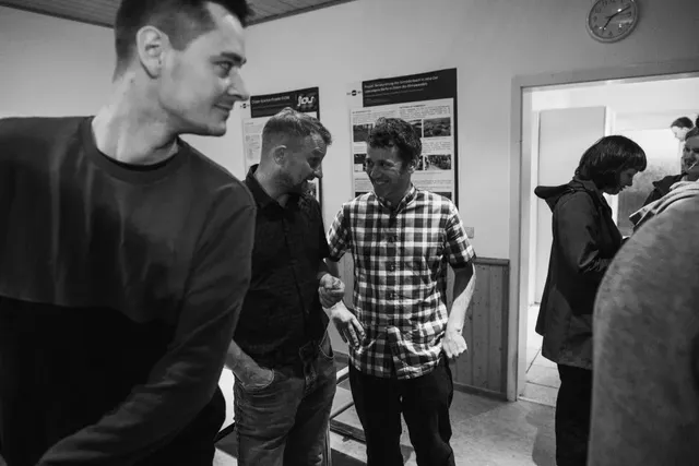 Drei Männer unterhalten sich in einem Raum, während im Hintergrund weitere Personen stehen; das Bild ist in Schwarz-Weiß gehalten.