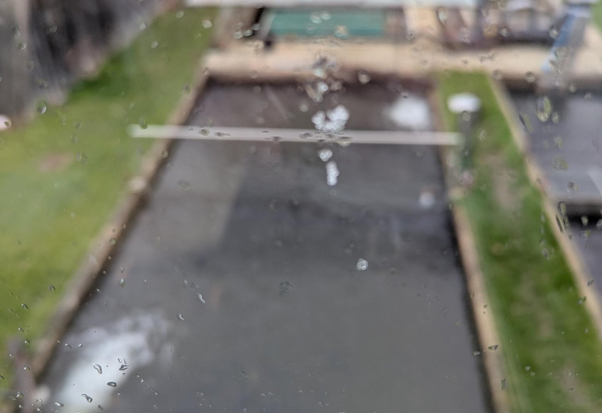 Blick durch ein Fenster mit Regentropfen auf mehrere rechteckige Außenbecken und eine überdachte Anlage im Hintergrund.